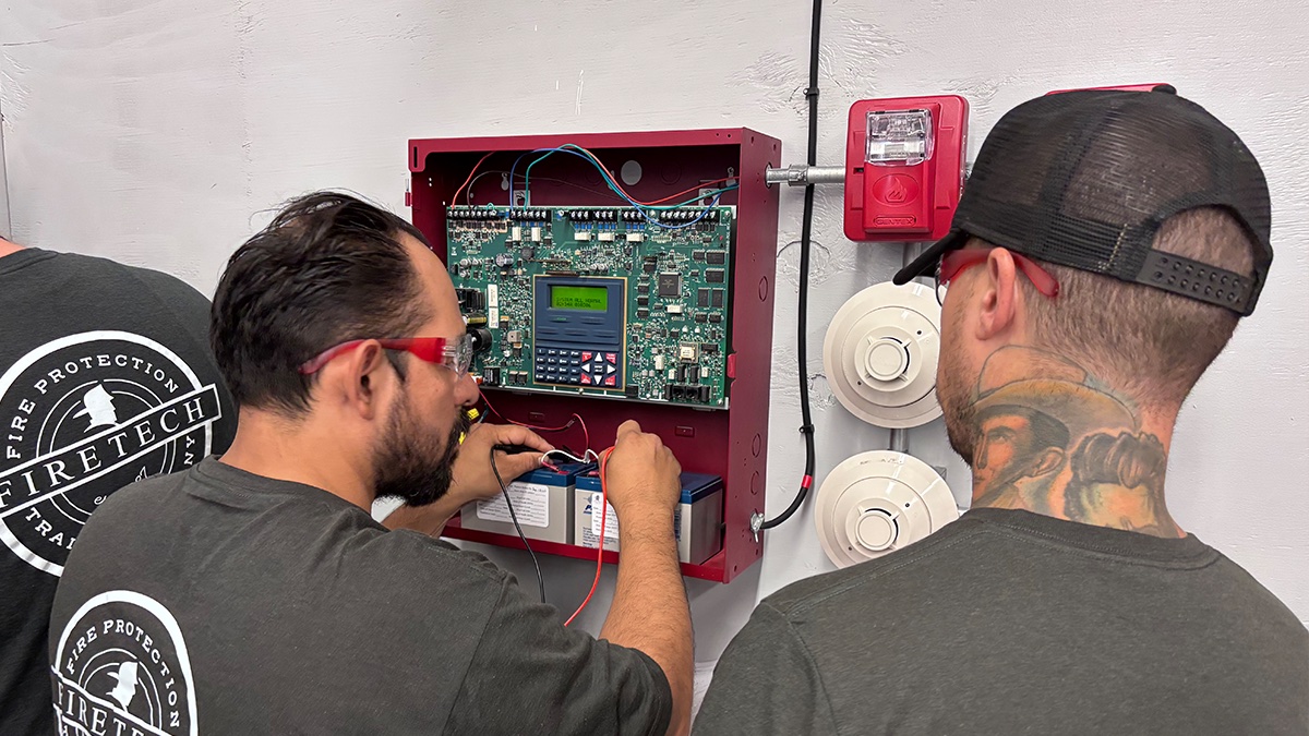 Two Fire Tech students wiring a fire alarm control panel during a hands-on workshop