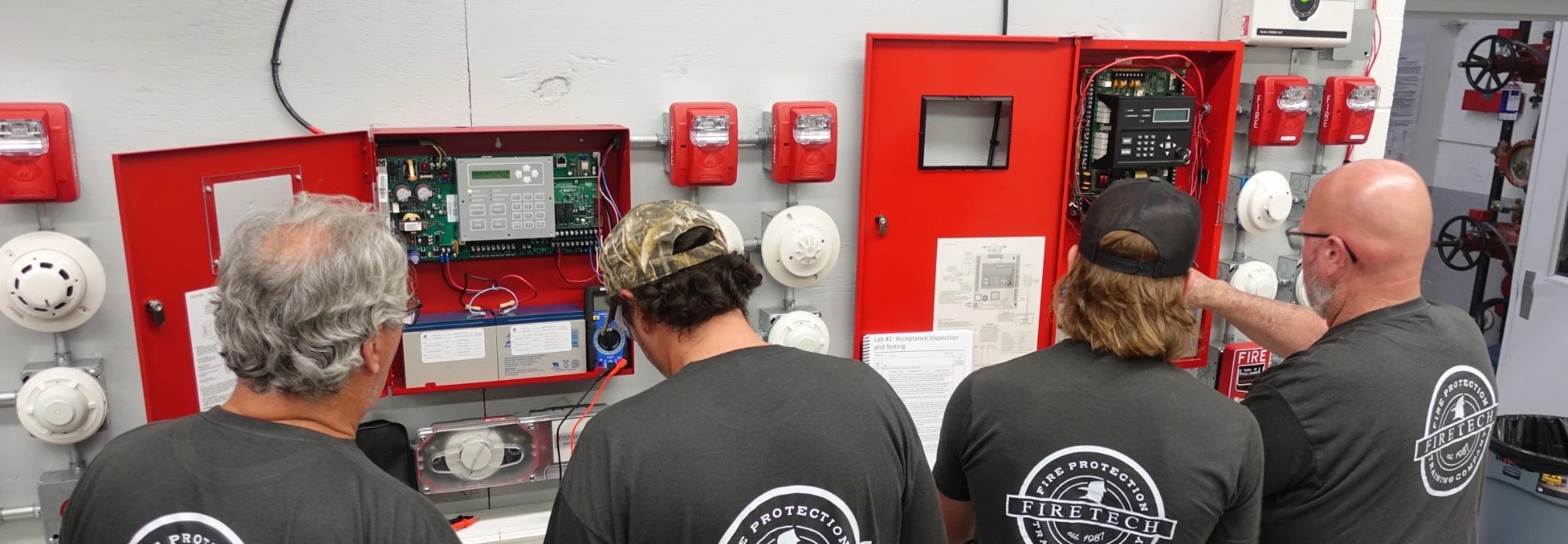 Four Fire Tech workshop attendees in Fire Tech shirts working together at a fire alarm training wall