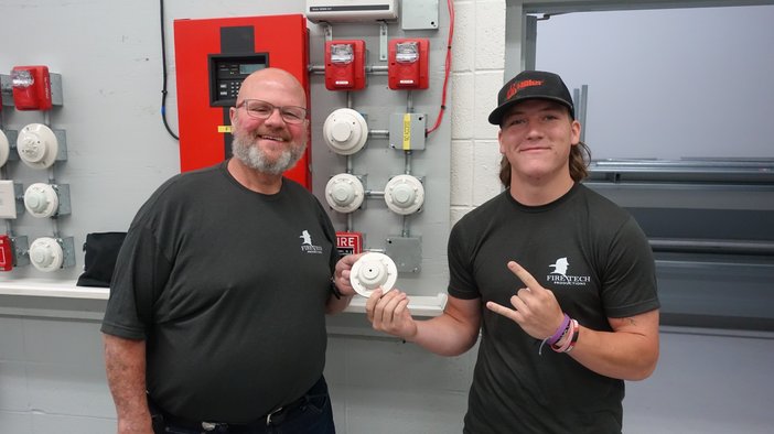 Two smiling Fire Tech workshop attendees holding a smoke detector in front of the alarm training wall